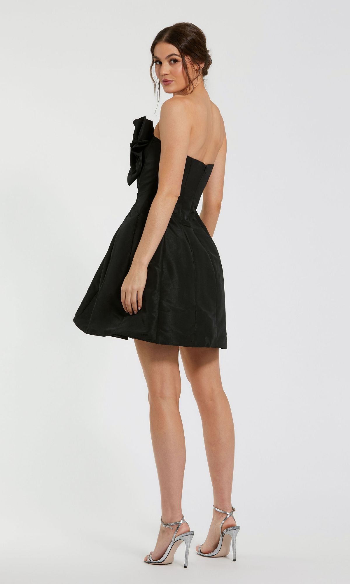 A woman models the Mac Duggal 49210 Strapless Short Bow Party Dress with silver high-heeled sandals, standing slightly turned from the camera against a plain background.